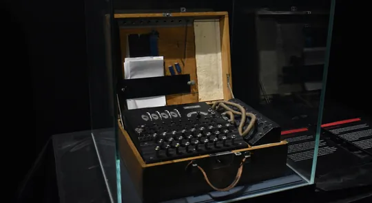 Enigma cipher machine on display behind glass: lampboard, rotor set, and paper tape inside an open wooden case